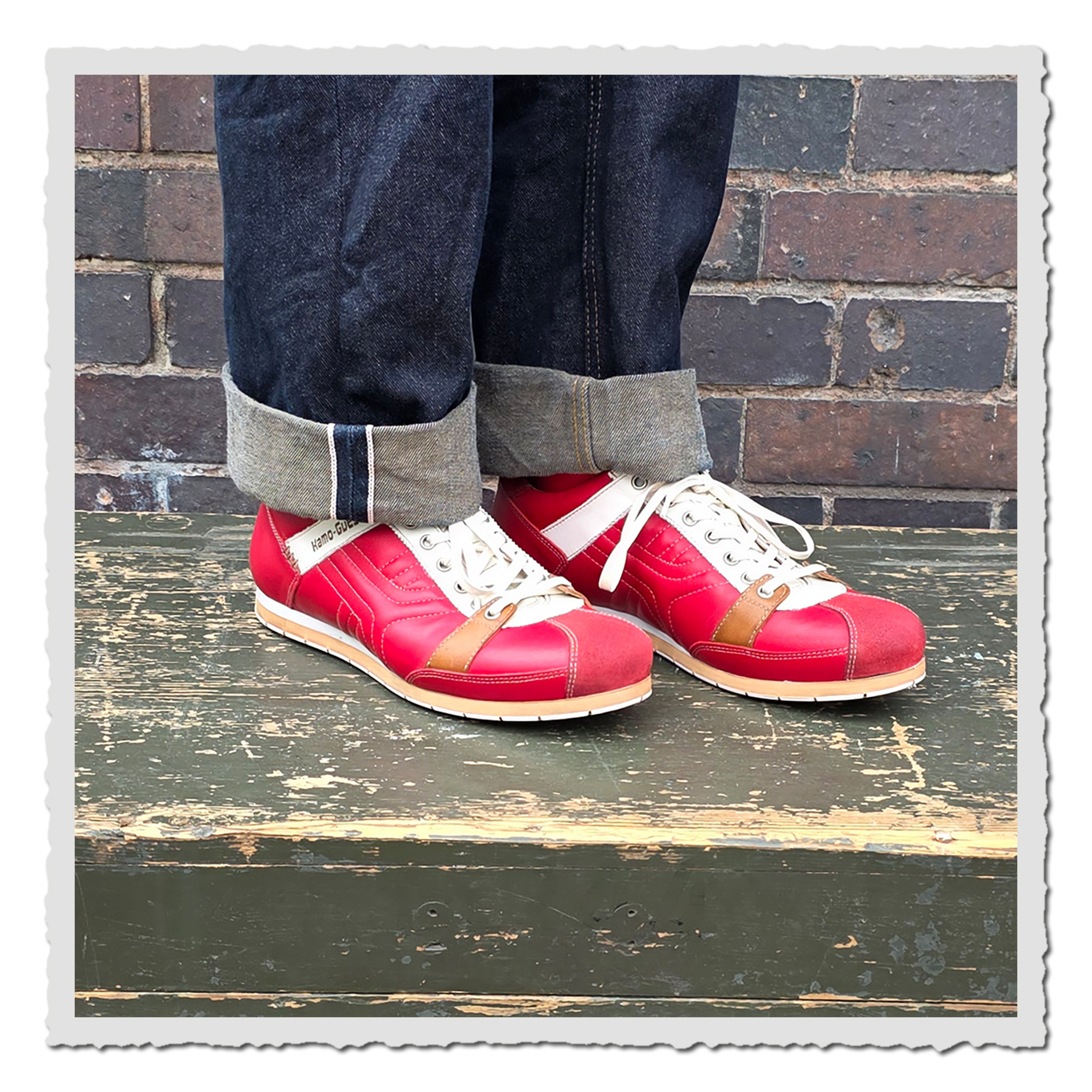 Rote Schnürschuhe an einer Person mit umgeschlagenen dunklen Jeans, auf grün lackierter Holzfläche vor Backsteinwand.